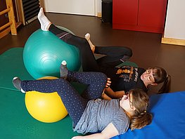 Mädchen und Therapeutin liegen auf dem Rücken, die Beine hat jeder auf einem Gymnastikball